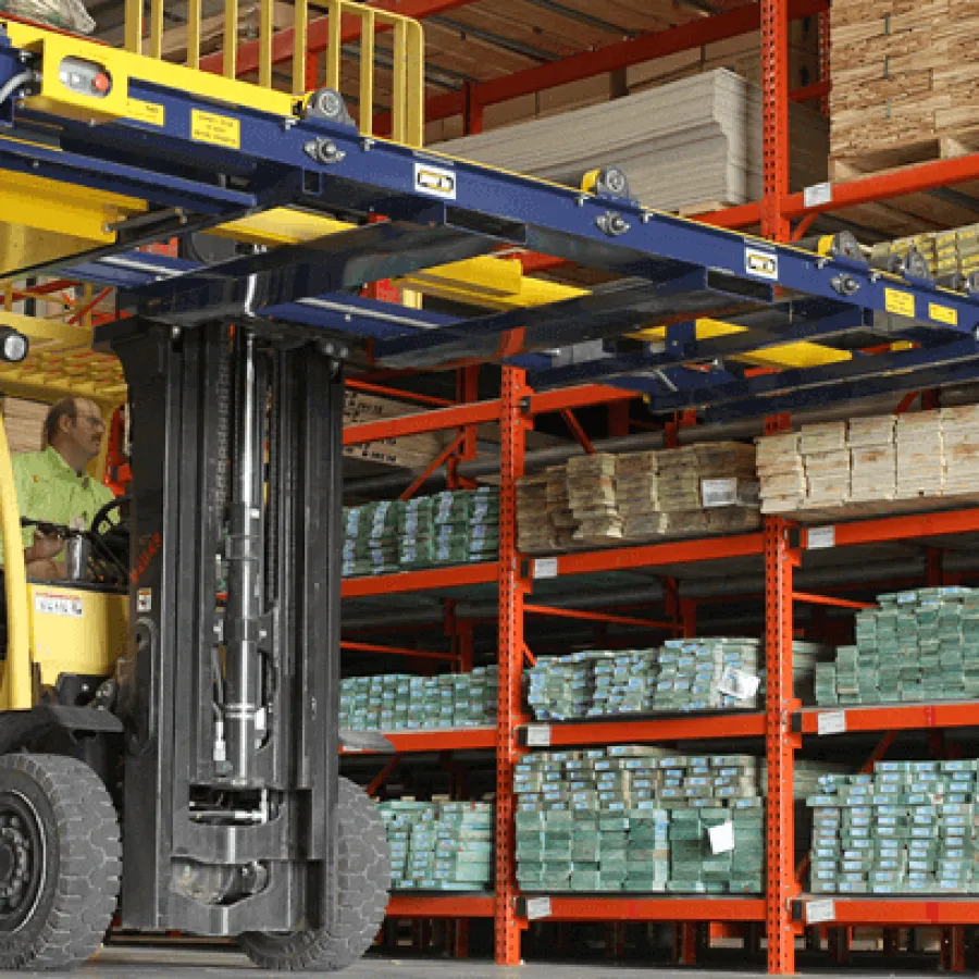 Forklift operator moving stacked materials in a large warehouse with orange metal shelving and wooden pallets.
