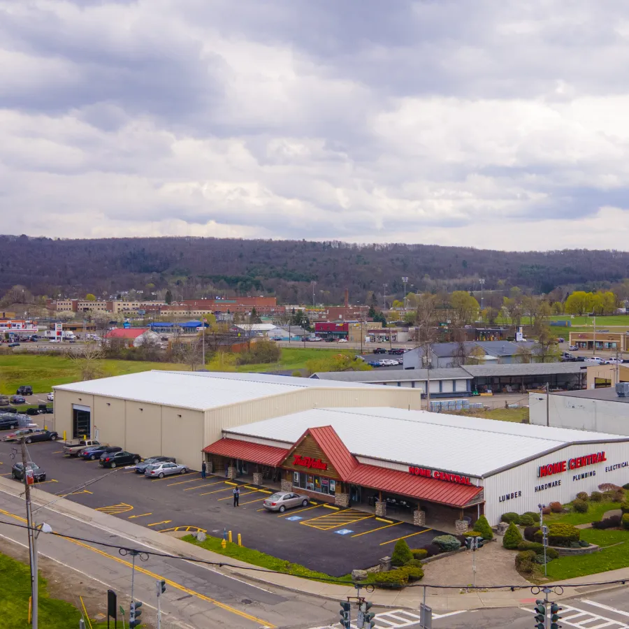 Aerial view of a commercial hardware and lumber store with parking lot and surrounding businesses under cloudy sky.