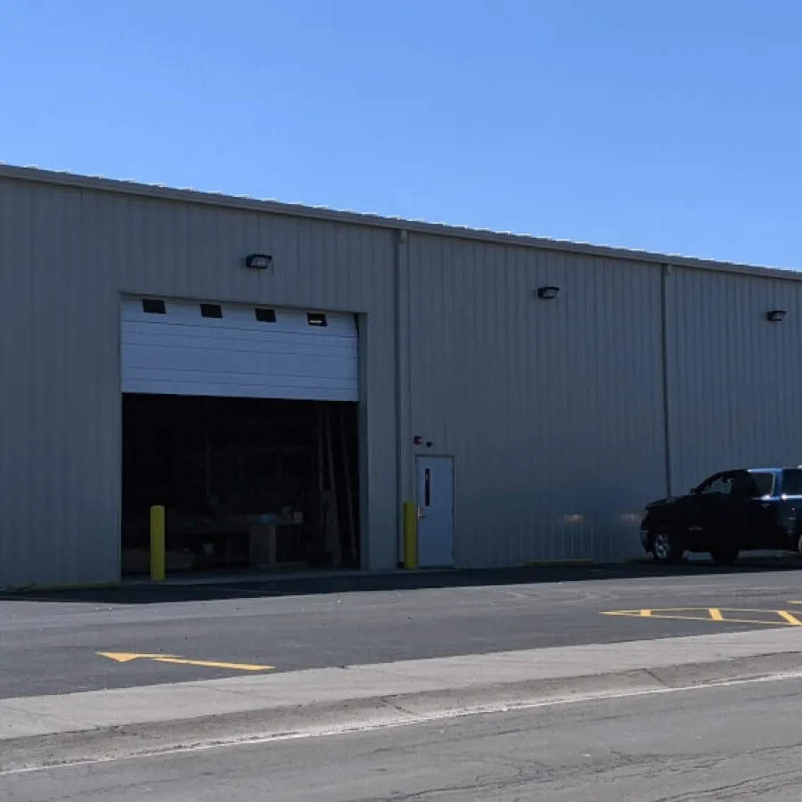 Gray industrial warehouse with open garage door and parked vehicles under clear blue sky.