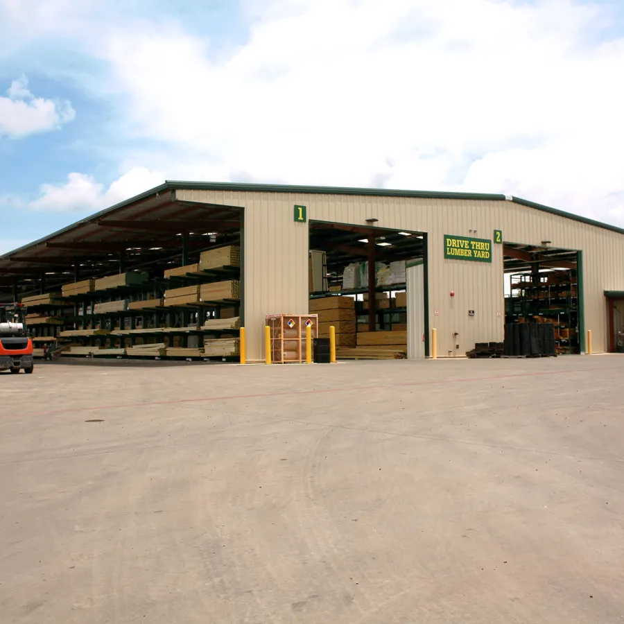Large lumber yard building with stacked wood and forklift under partly cloudy sky.