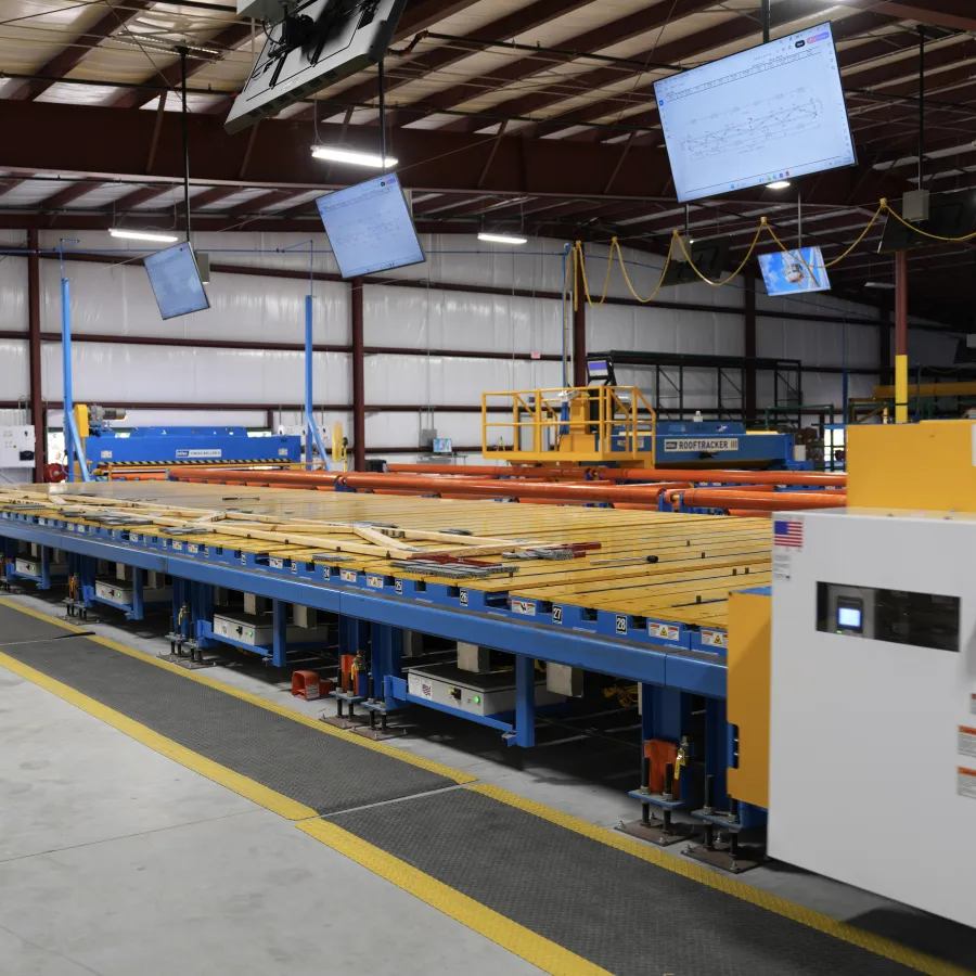 Long conveyor belt transporting wooden planks inside a spacious factory with overhead screens and industrial equipment.