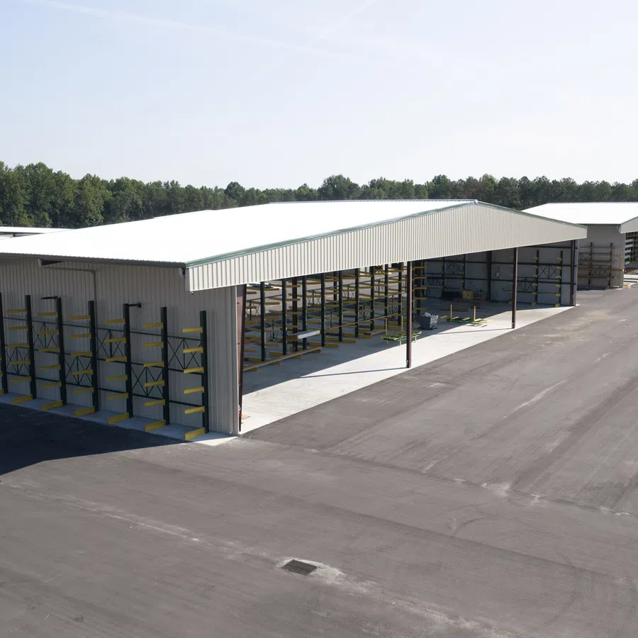 Two large metal storage sheds with open sides and cantilever rack shelving on a wide paved lot under clear sky