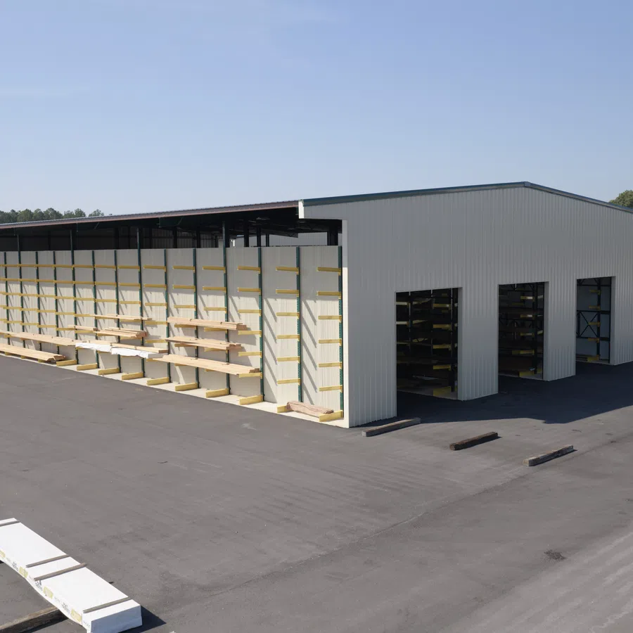 Large outdoor storage facility with metal building and wood planks stored on exterior racks under clear sky