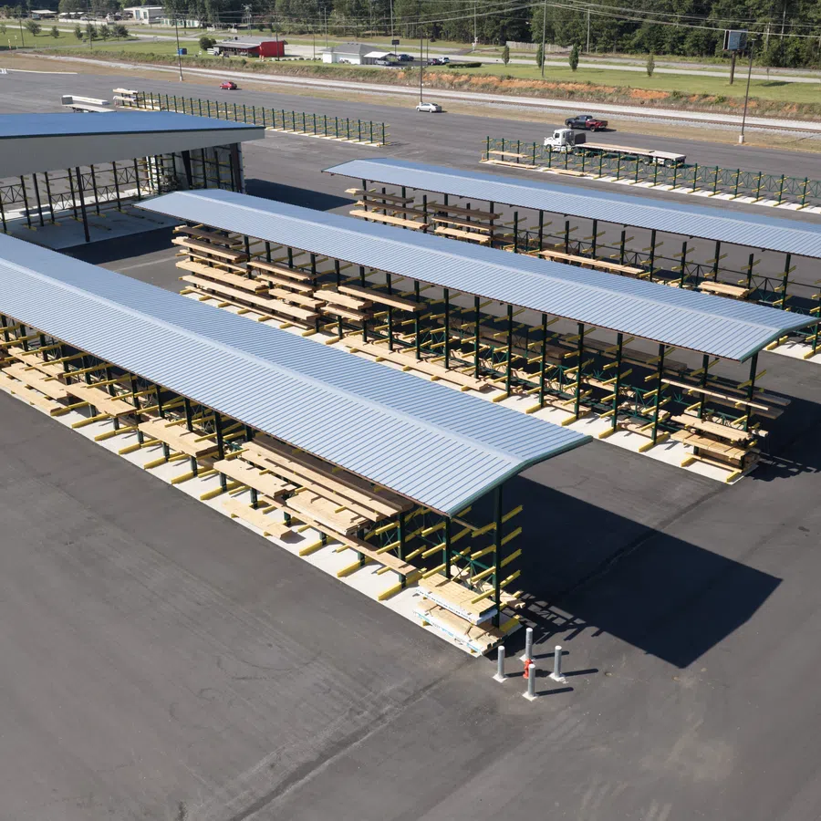 Large outdoor lumber storage racks with metal roofs in a paved yard beside a road and greenery in the background