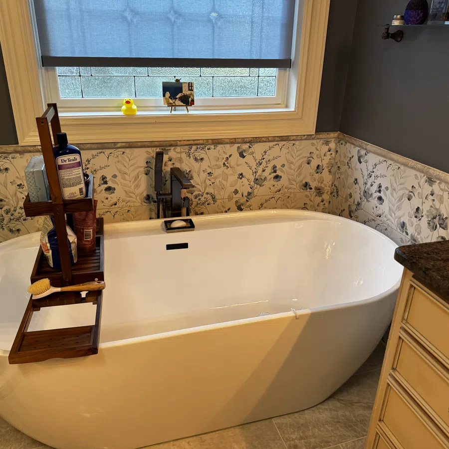Modern white freestanding bathtub with wooden caddy, floral wallpaper, blue window shade, and granite countertop.