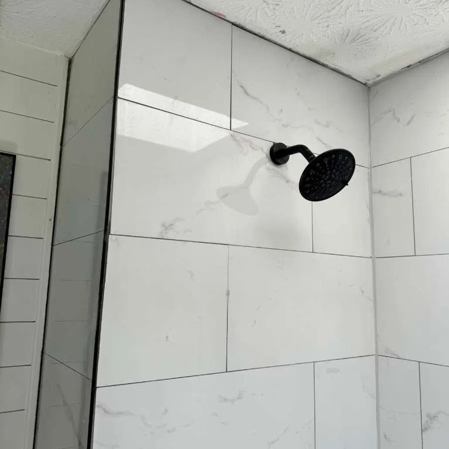 Modern shower with black showerhead and white marble-style tiles on walls in a bathroom corner.