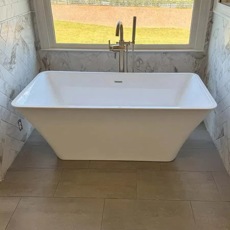White freestanding bathtub with modern faucet under a large window overlooking a grassy yard.