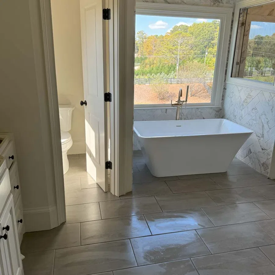 Modern bathroom with a freestanding bathtub, large window with outdoor view, and adjacent toilet room.