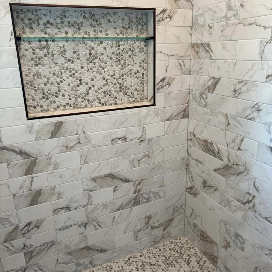 Shower corner with marble subway tiles and pebble mosaic floor and niche with glass shelf.