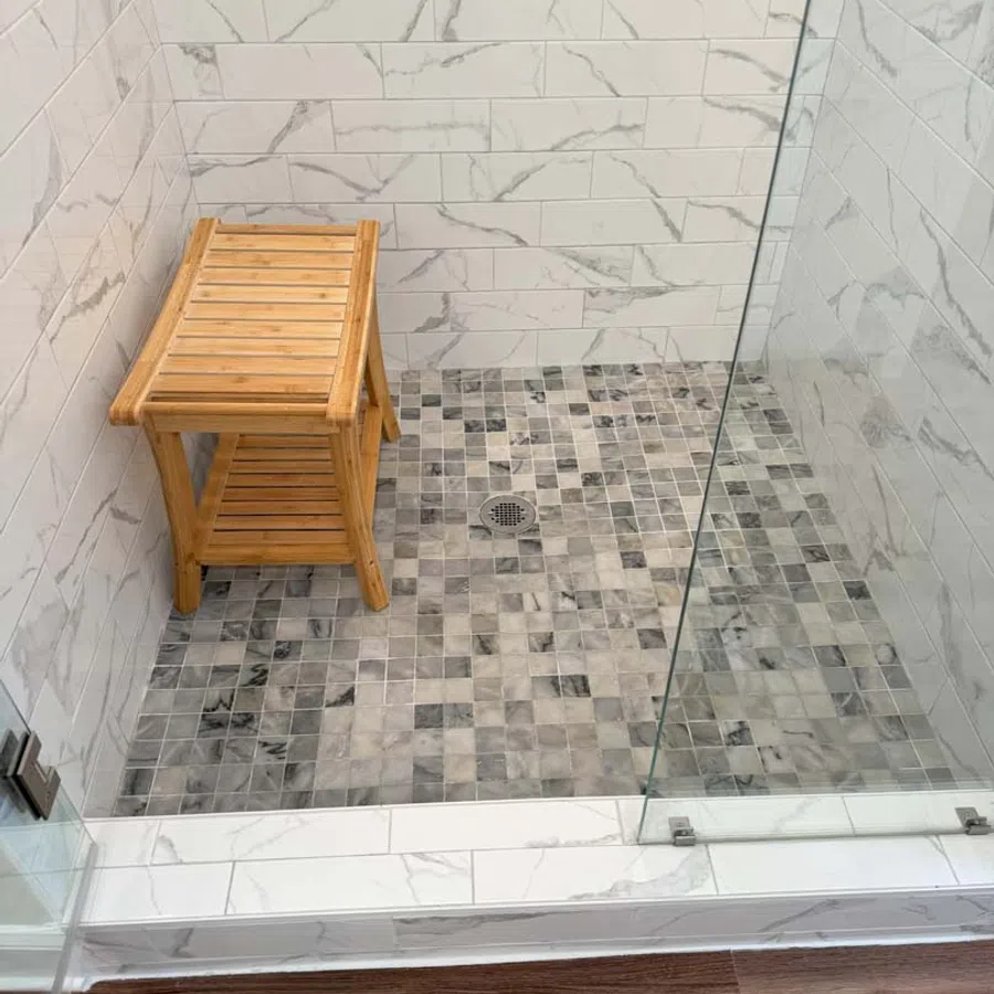 Modern walk-in shower with glass door, mosaic tile floor, marble wall tiles, and wooden shower stool.