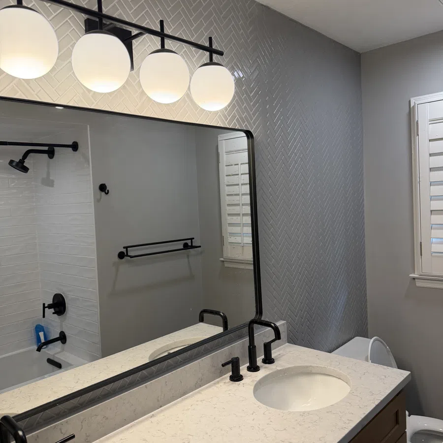 Modern bathroom with double sinks, black fixtures, large mirror, marble countertop, and herringbone tile wall.