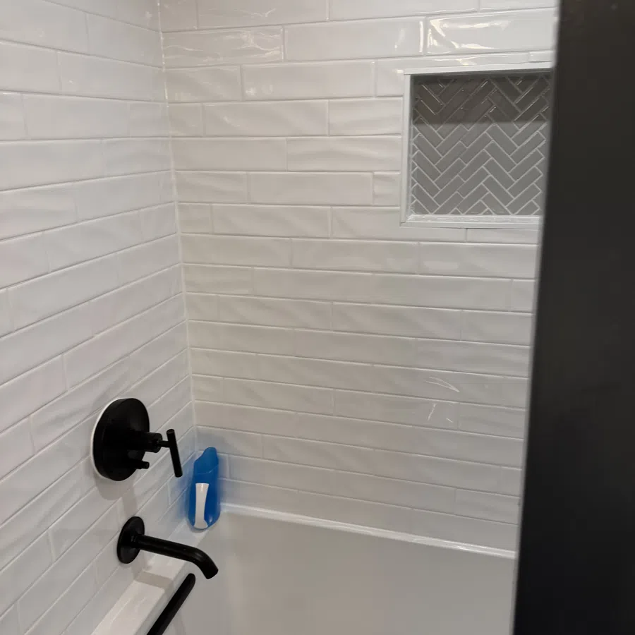 White tiled bathroom shower with black faucet fixtures and a blue shampoo bottle on the tub edge.
