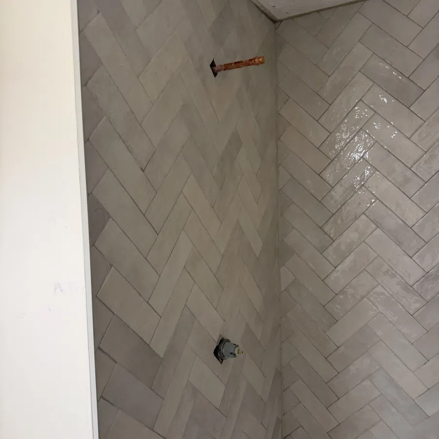 Gray herringbone tile wall in a corner with exposed plumbing pipes for a shower installation.
