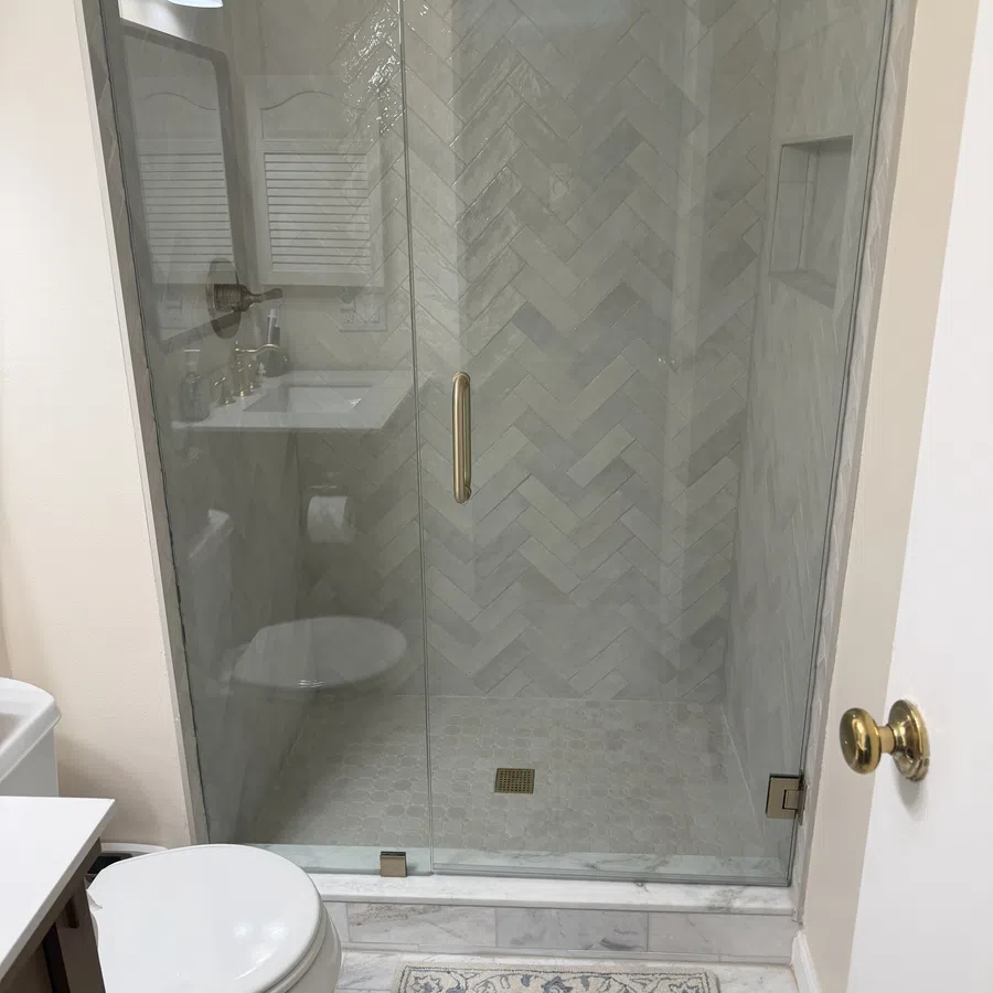 Modern bathroom with glass shower door, herringbone tile pattern, white toilet, and decorative rug on marble floor.