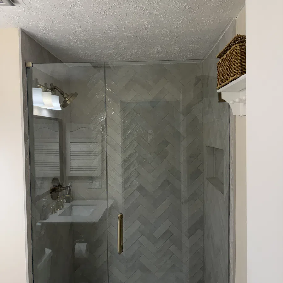 Modern bathroom shower with glass door, herringbone tile pattern, and brass handle, featuring a wicker basket shelf.