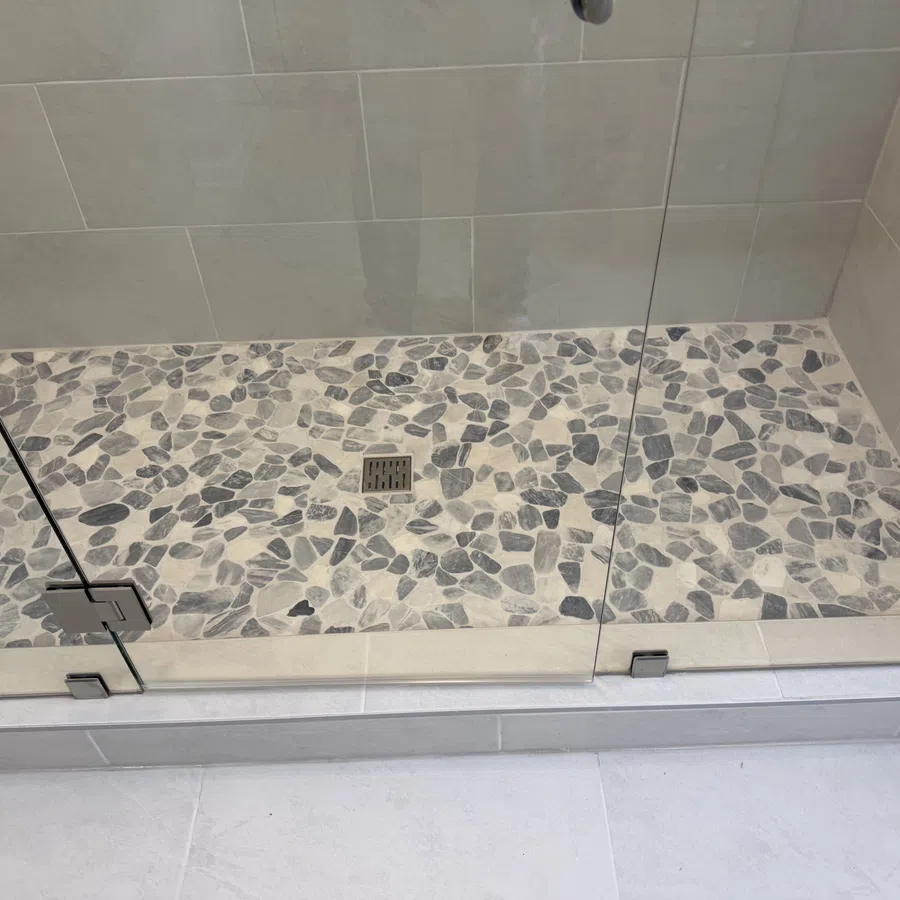 Modern shower area with pebble stone floor tiles and glass door enclosure with metal hinges and drain.