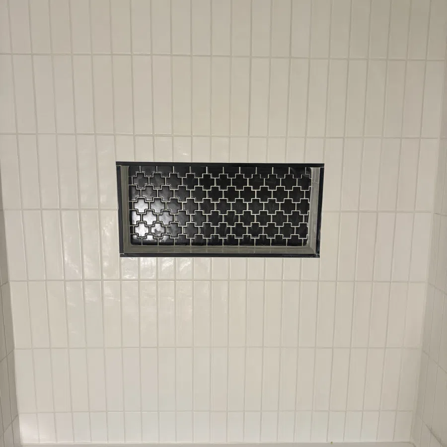 White tiled bathroom wall with a decorative black vent grate above a white bathtub