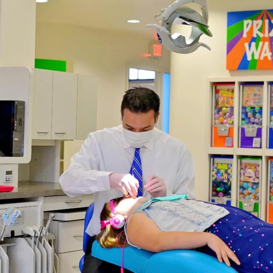 Dentist treating a young girl wearing pink headphones in a bright pediatric dental clinic with a colorful prize wall.