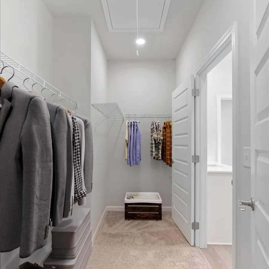 Bright walk-in closet with hanging clothes, storage boxes, carpeted floor, and a wooden storage bench at the end.