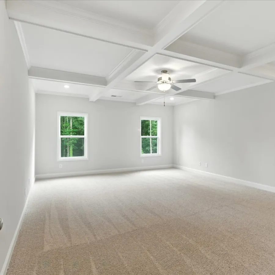 Spacious empty room with beige carpet, white walls, coffered ceiling, ceiling fan, and two windows showing greenery.