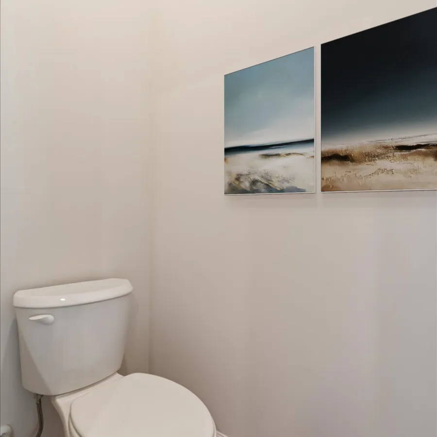 White toilet in a minimalist bathroom corner with two abstract landscape paintings on the wall above wooden floor.