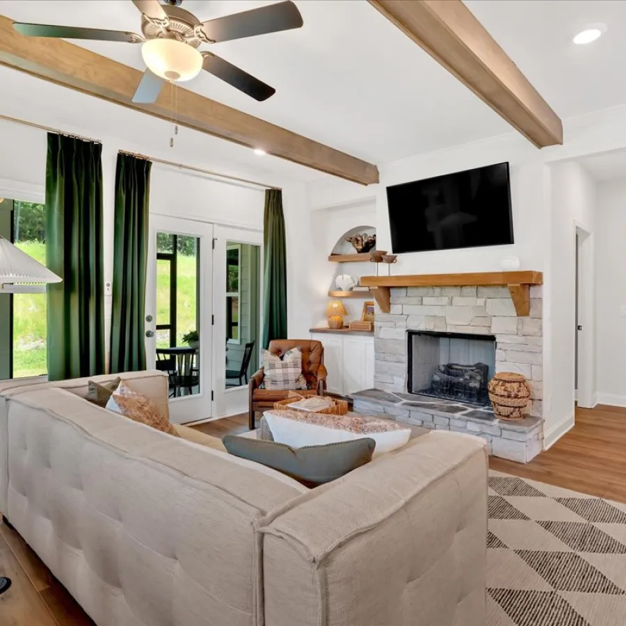 Cozy living room with beige sofa, stone fireplace, wood beams, ceiling fan, and large windows with green curtains.