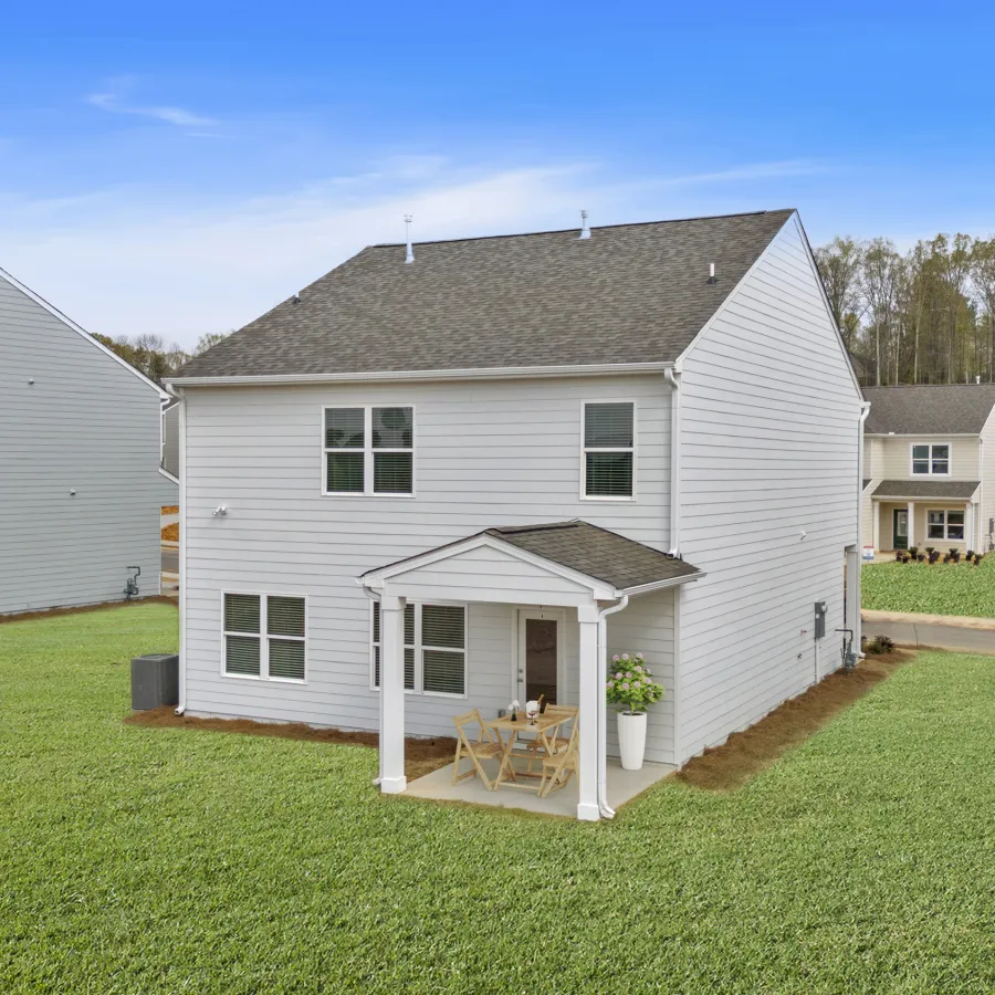 White two-story house with covered patio, green lawn, neighboring homes, and wooded background under blue sky.