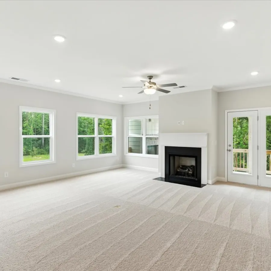 Bright empty living room with beige carpet, ceiling fan, fireplace, multiple windows, and glass door to outside deck.