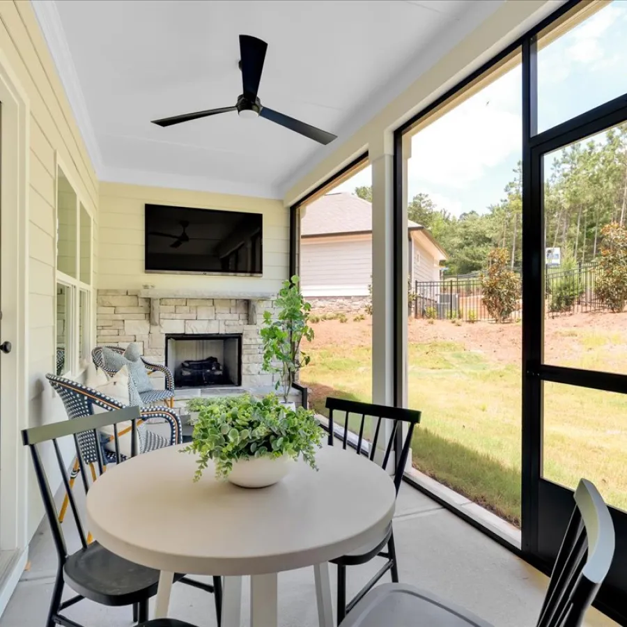 Cozy screened porch with round table, black chairs, ceiling fan, fireplace, and greenery overlooking backyard.