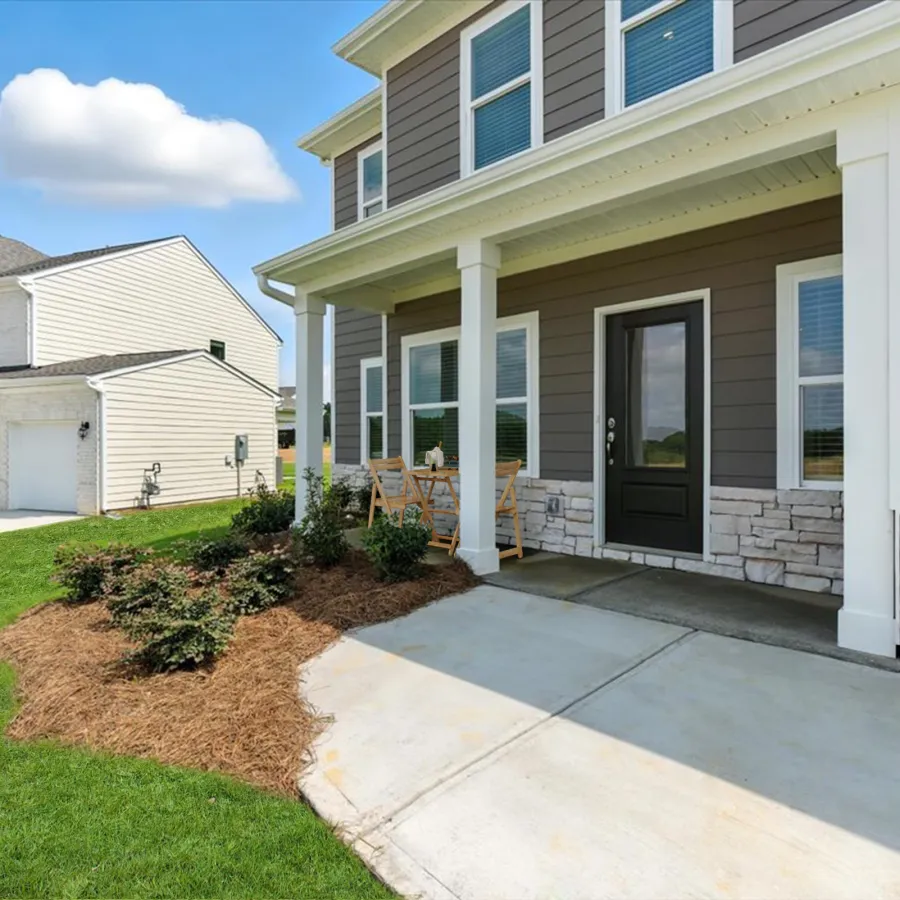 Front porch of modern two-story house with patio chairs, black door, stone accents, and landscaped lawn under blue sky.