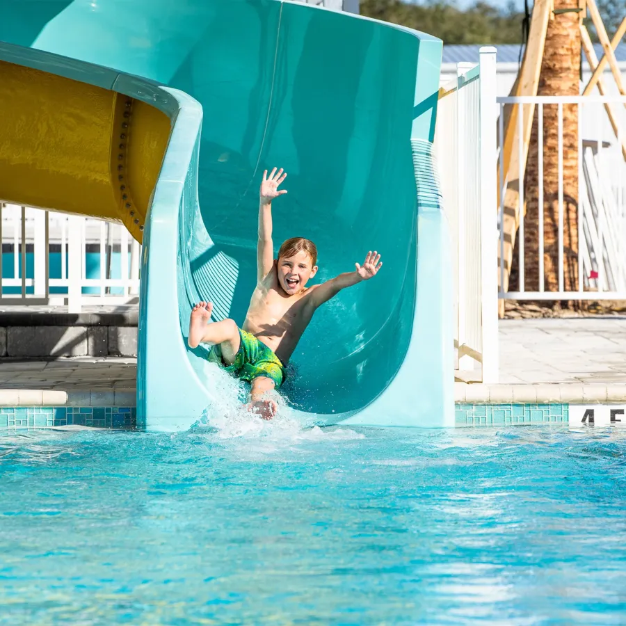 Excited boy in green swim trunks sliding down blue water slide splashing into pool on sunny day