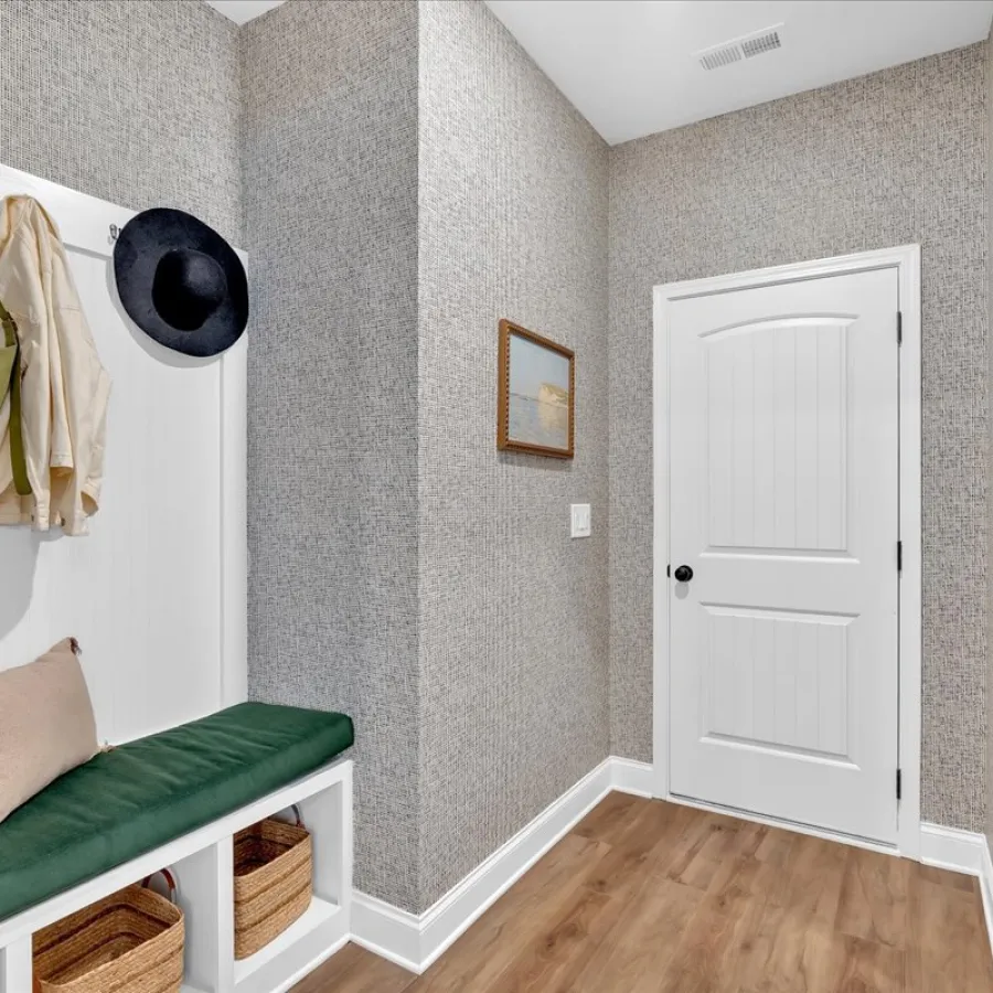 Modern entryway with cushioned bench, hanging coats and hats, wooden floor, and white paneled door.