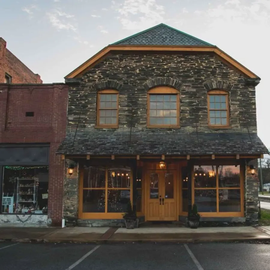Rustic stone and brick storefronts with large windows and wooden doors at dusk in a small town street.