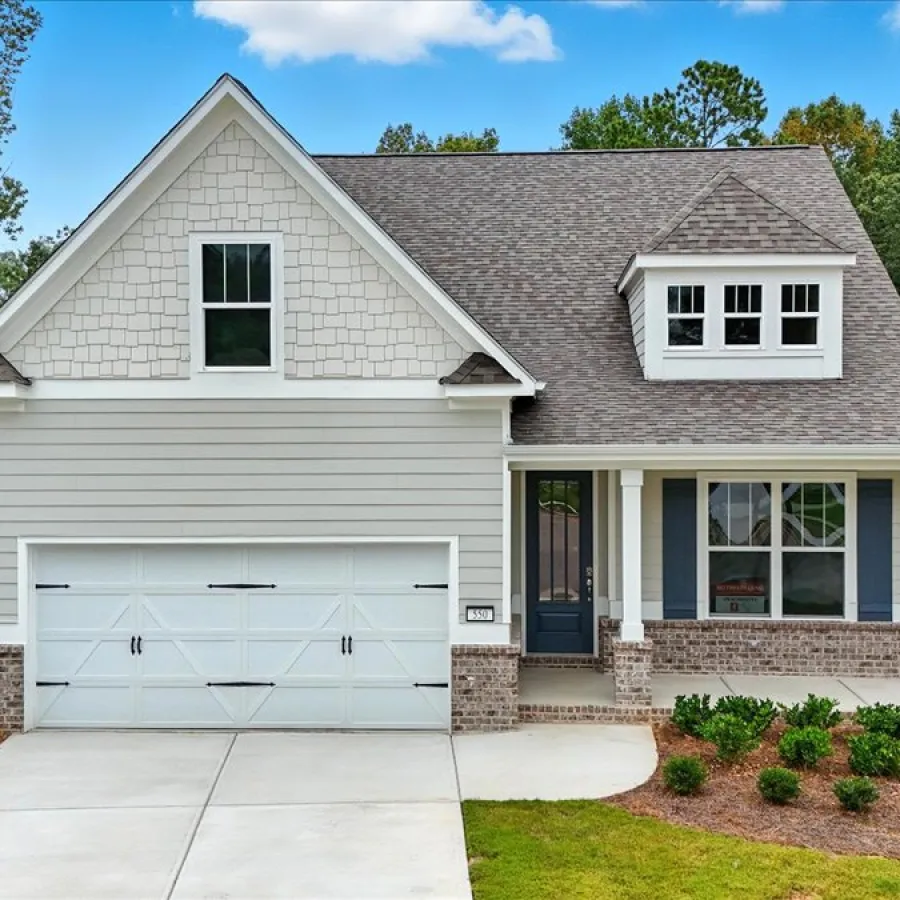 Modern two-story gray house with a white garage door, front porch, and a landscaped yard surrounded by trees.
