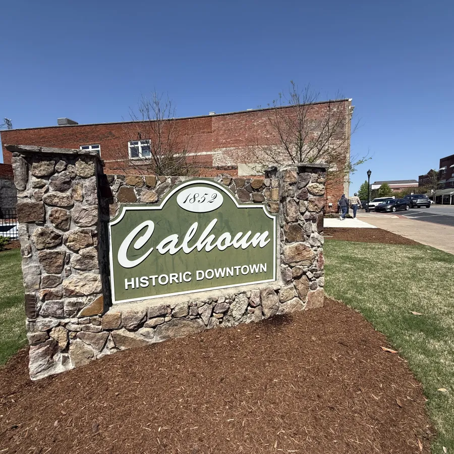 Stone sign announcing Historic Downtown Calhoun with blue sky and nearby buildings in the background.