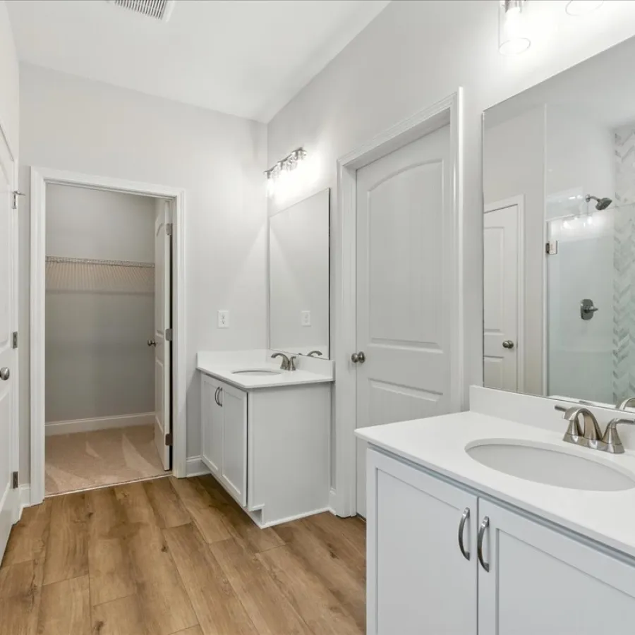 Modern bathroom with white cabinetry, dual sinks, large mirror, glass shower, and wood-style flooring.