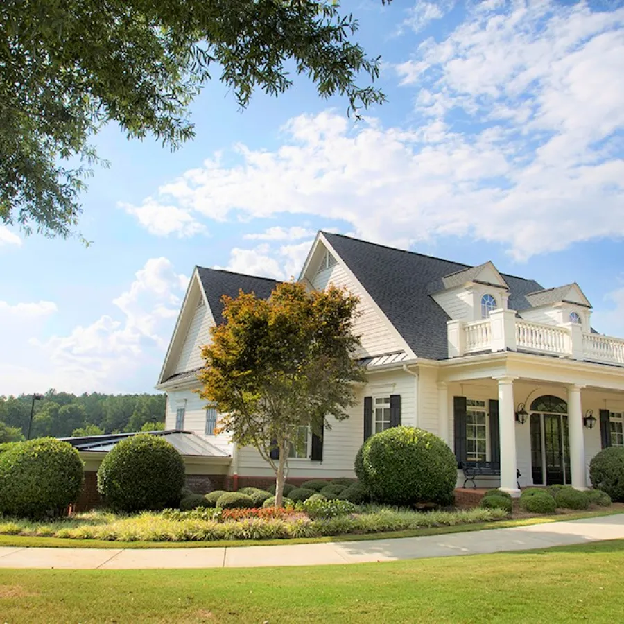 White two-story house with black shutters surrounded by manicured bushes and trees under a blue sky with clouds.
