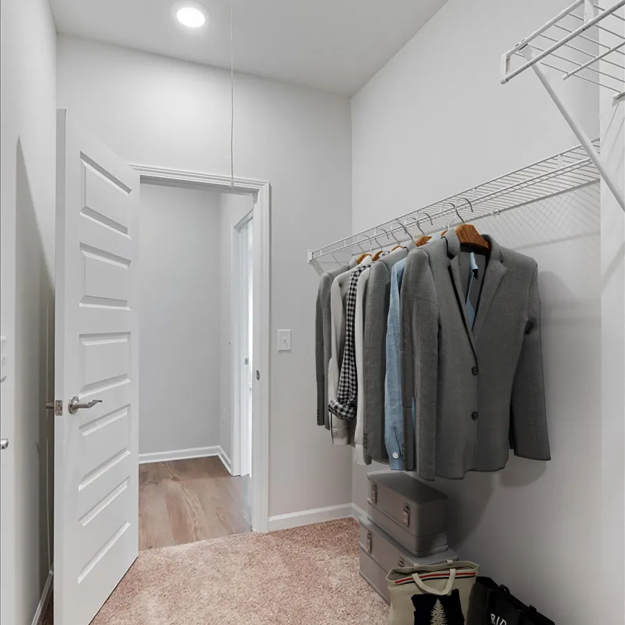 Small walk-in closet with hanging jackets, carpeted floor, overhead light, door open to wood floor hallway.