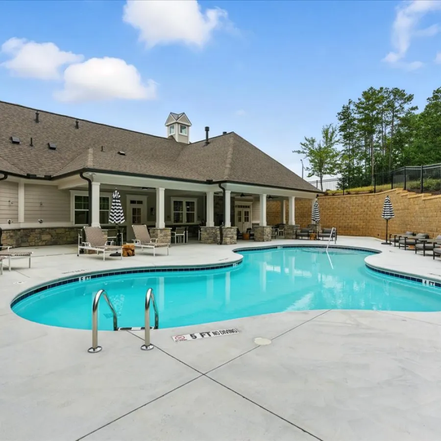 Outdoor residential swimming pool with lounge chairs, umbrellas, and a covered patio under a blue sky.