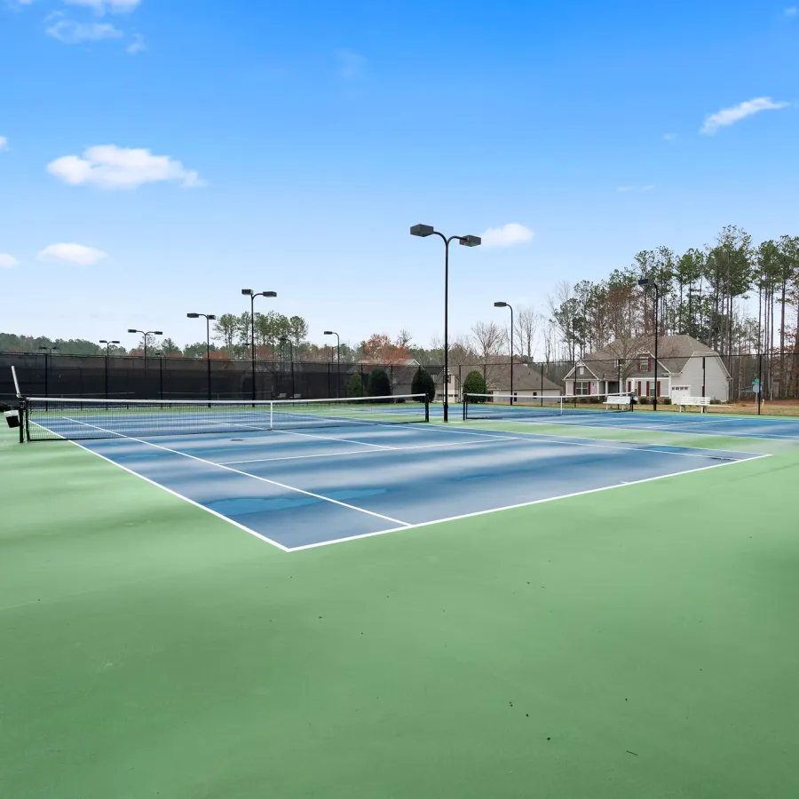 Outdoor tennis courts with blue playing surface and green perimeter under a clear blue sky with surrounding trees and homes.