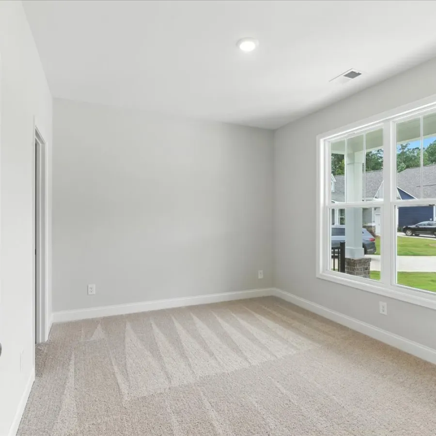 Empty bright room with beige carpet, light gray walls, white trim, and a large window overlooking houses outside.