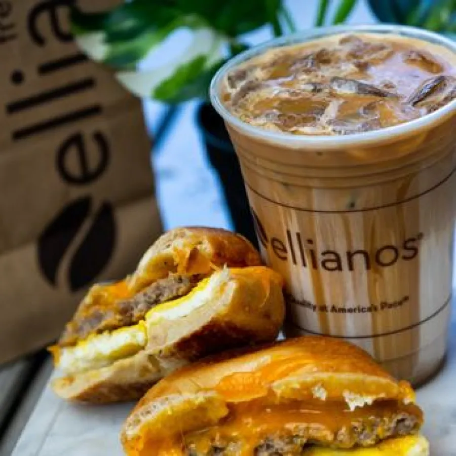 Iced coffee beside a breakfast sandwich with egg, sausage, and melted cheese on a white surface.