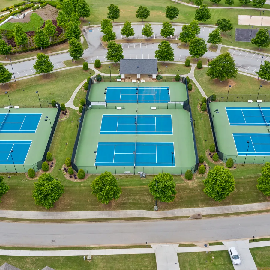 Aerial view of three fenced blue and green tennis courts surrounded by trees and a nearby parking lot.