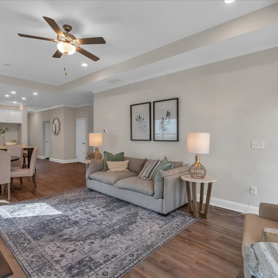 Modern living room with gray sofa, armchair, patterned rug, ceiling fan, and open dining area with hardwood floors.