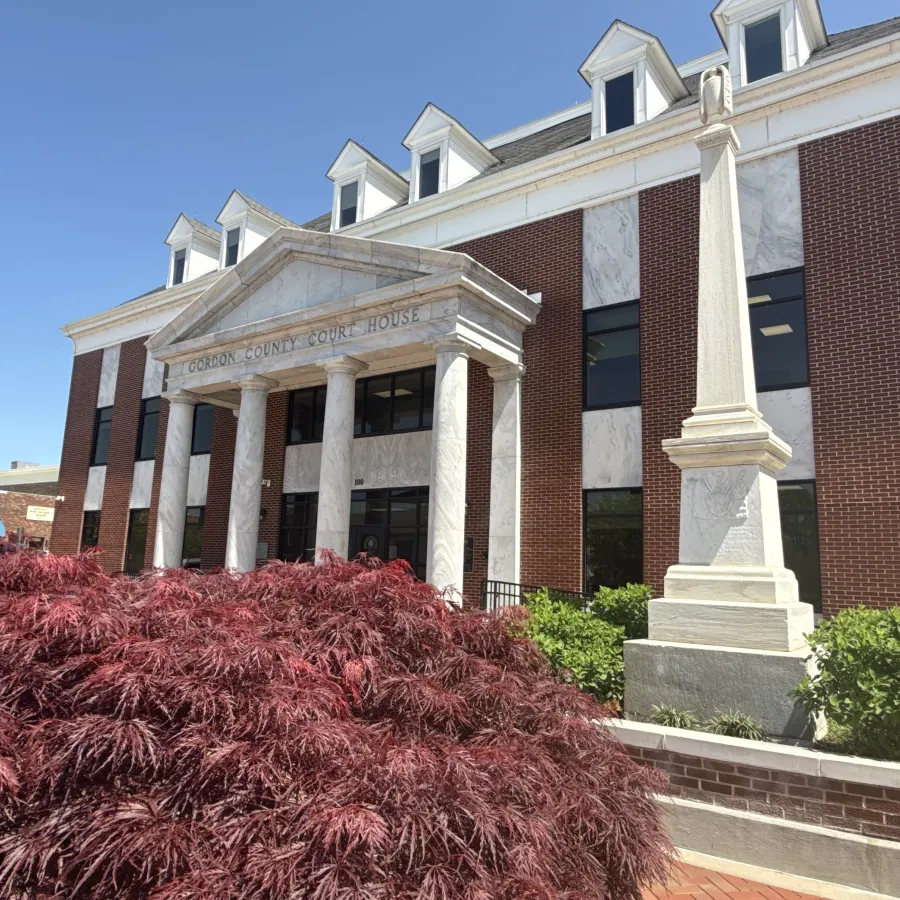 Gordon County Courthouse with white columns, red brick facade, obelisk monument, and red foliage under clear sky.