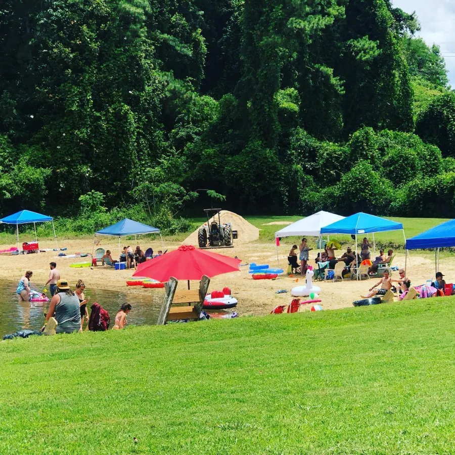 People enjoying a sunny day at a small sandy beach with colorful umbrellas and canopies by a lake.