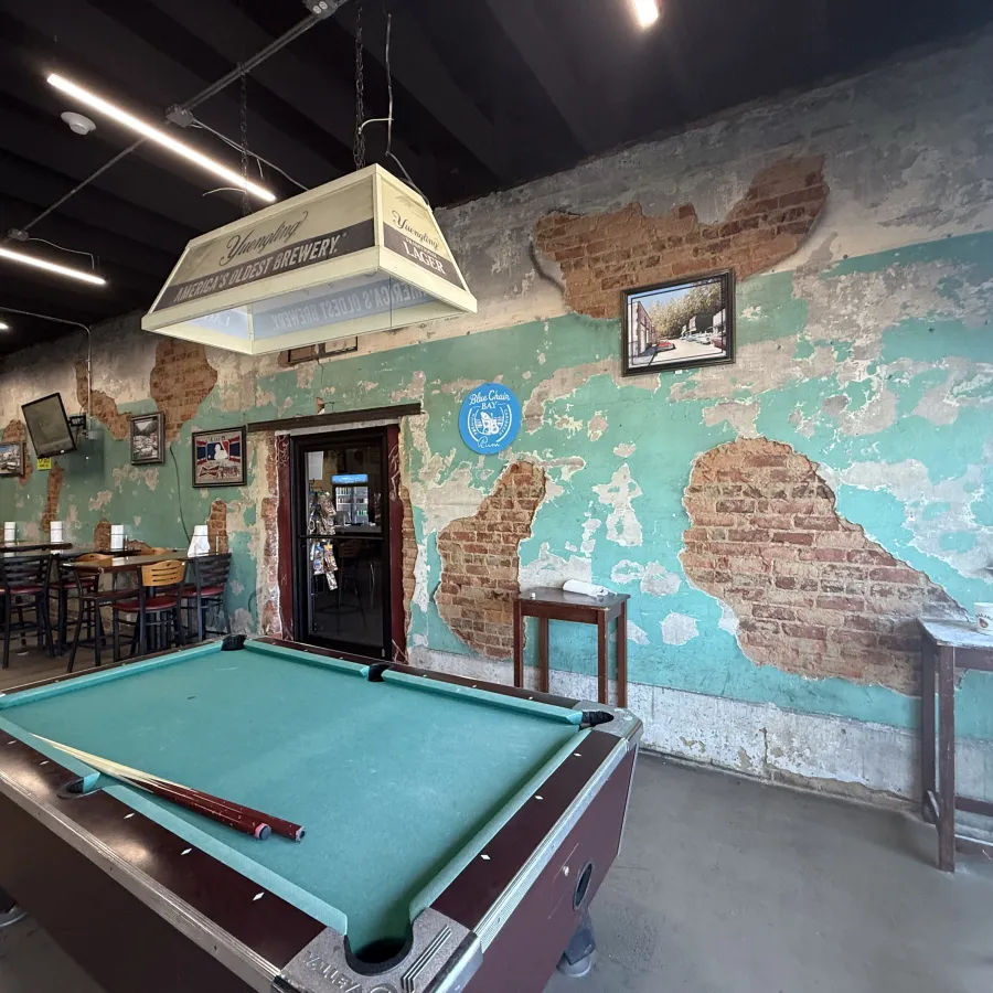 Pool table with cues in a rustic bar featuring peeling turquoise paint and exposed brick walls.