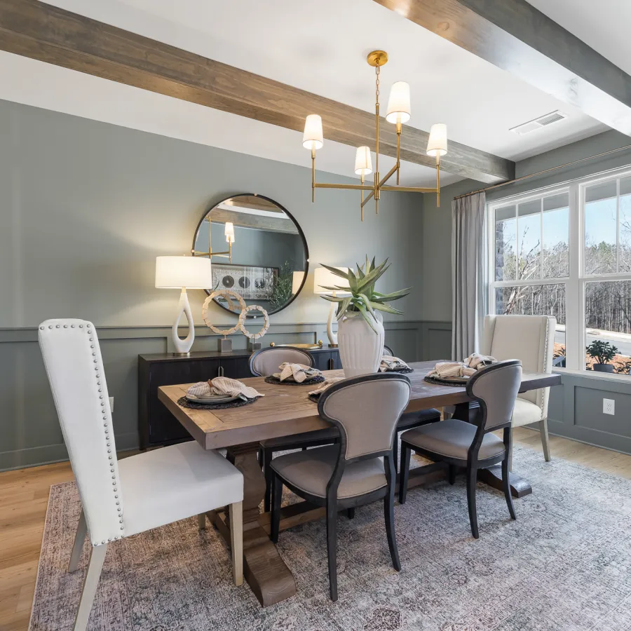 Modern dining room with wooden table, mixed chairs, round mirror, chandelier, large window, and potted plants.