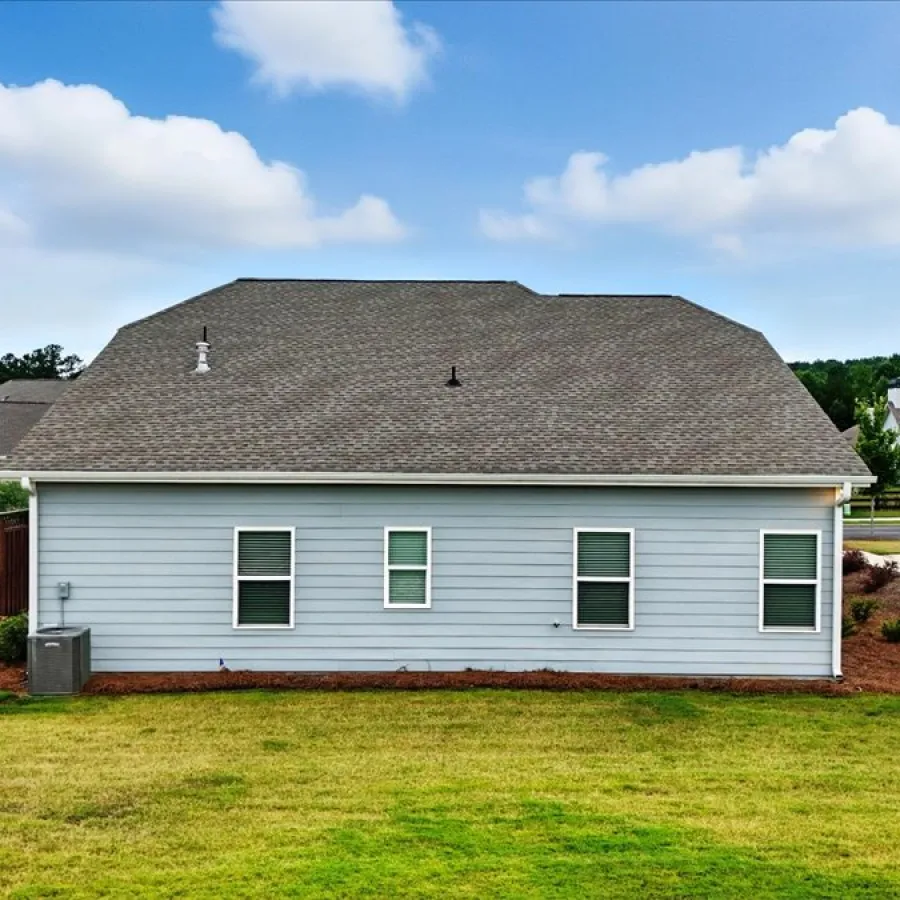Suburban house with gray siding, four windows, a shingled roof, and a green lawn under a partly cloudy sky.
