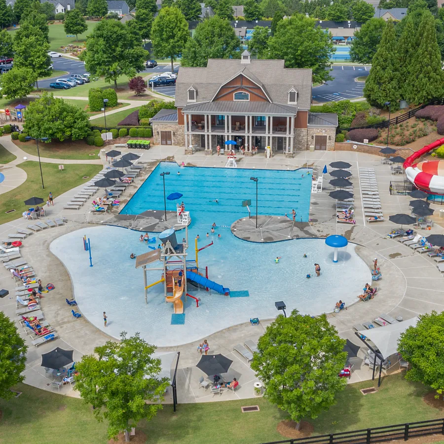 Aerial view of a community pool area with water slides, splash pads, lounge chairs, and a clubhouse surrounded by greenery.